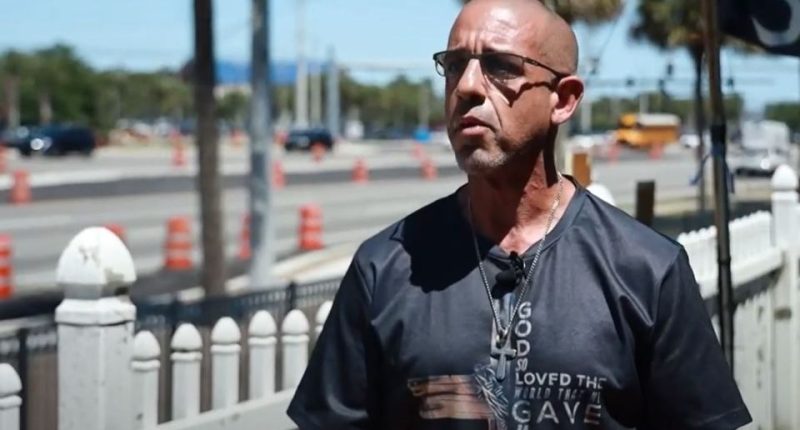 Man in glasses and a religious t-shirt speaks outdoors.