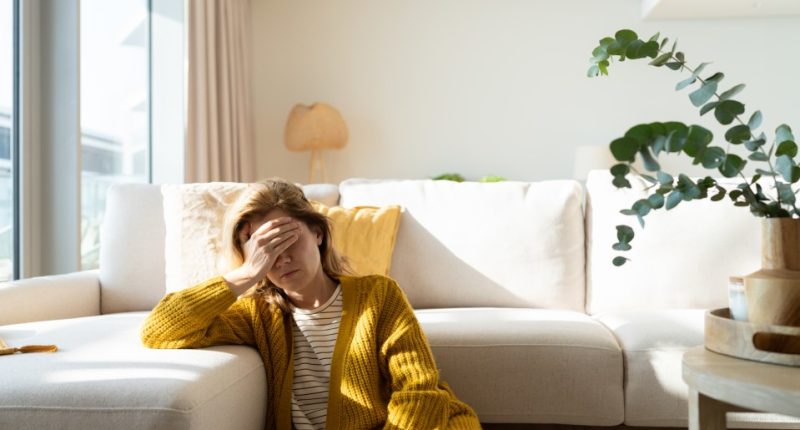 Woman sitting on couch holding her head, experiencing a headache.