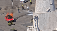 A burning car being extinguished by firefighters near a monument.