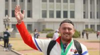 A man in North Korea wearing a red tracksuit and a gold medal waves.