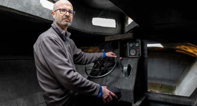 Man sitting at the controls of a small, dark submarine.