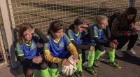 Girls' soccer team from Kherson, Ukraine, sitting near a fence before a match.