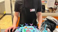 A young man in a cafe apron smiles at the camera, in front of a mermaid-themed first anniversary cupcake.