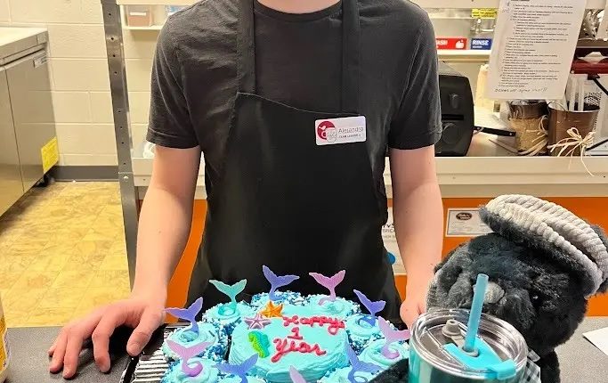 A young man in a cafe apron smiles at the camera, in front of a mermaid-themed first anniversary cupcake.