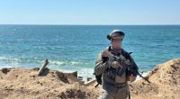 IDF reservist Captain U standing by the ocean with a rifle.