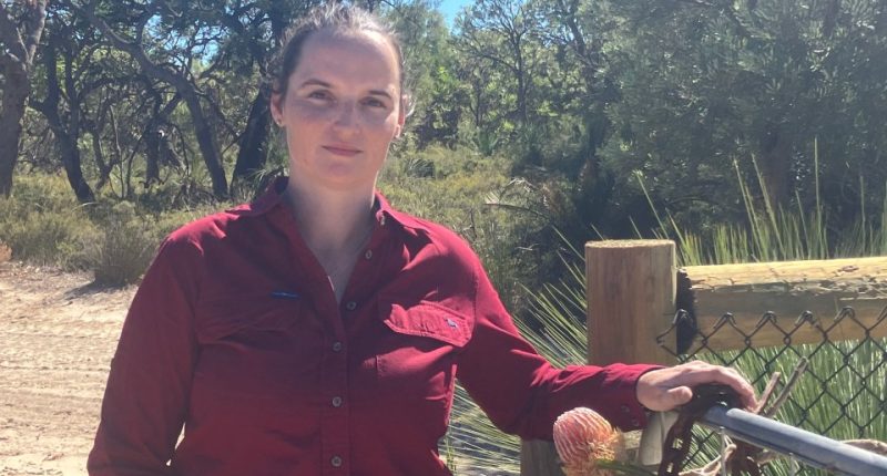 Woman in red shirt standing by a gate in a natural setting.