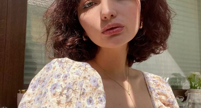 Portrait of a young woman with short, dark curly hair wearing a floral dress and gold necklace.