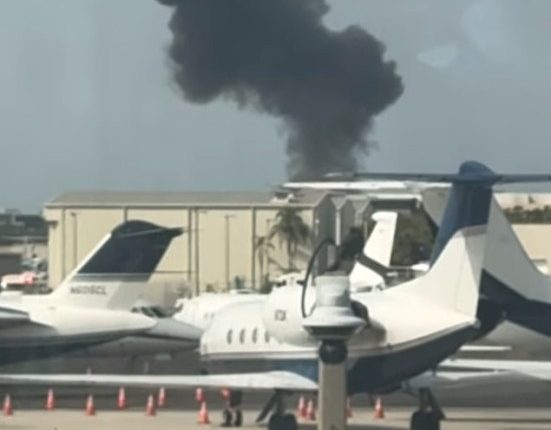 Black smoke plume over airport tarmac with private jets.
