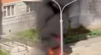Screenshot of a burning car emitting thick black smoke near a residential building.