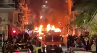 Burning vehicle in city street surrounded by onlookers.