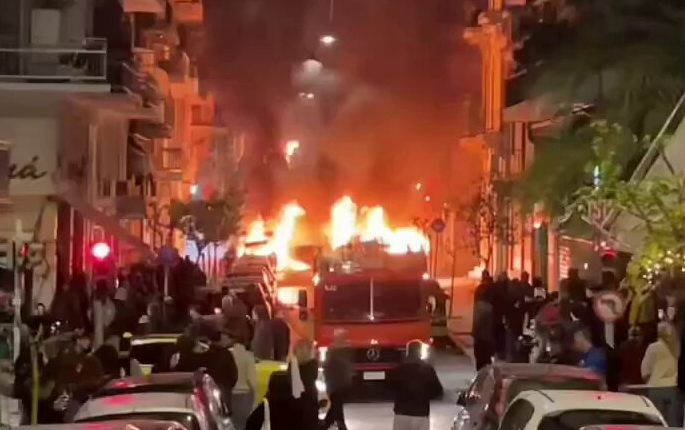 Burning vehicle in city street surrounded by onlookers.