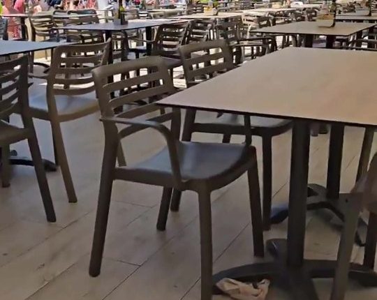 Outdoor restaurant patio with tables and chairs, overlooking a beach and city.