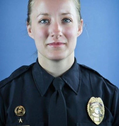 Headshot of a woman with blond hair pulled back, wearing a dark collared shirt against a blue background.