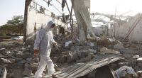 A person in protective gear walks through the rubble of a bombed-out building.