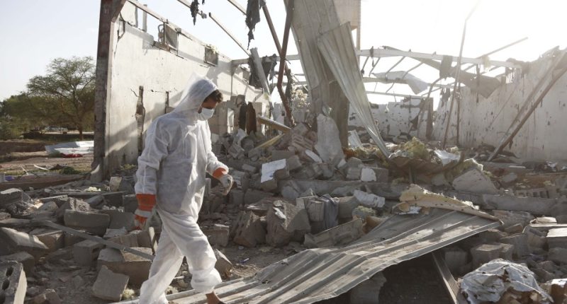 A person in protective gear walks through the rubble of a bombed-out building.