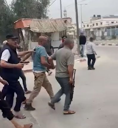 A group of armed men escorting a man in a light blue shirt down a street.