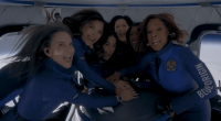 Six women in blue flight suits huddle together in a spacecraft.