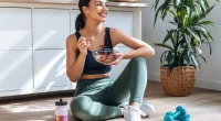 Shot of athletic woman eating a healthy bowl of muesli with fruit sitting on floor in the kitchen at home