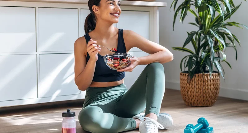 Shot of athletic woman eating a healthy bowl of muesli with fruit sitting on floor in the kitchen at home