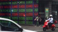 SYDNEY, AUSTRALIA - APRIL 08: Pedestrians are reflected in a window as they walk past an electronic stock board at the ASX Ltd. exchange centre on April 08, 2025 in Sydney, Australia. Australia is facing a baseline 10% tariff on all exports to the U.S., imposed under President Trump's "Liberation Day" trade measures, which have sparked widespread criticism from Australian leaders and industries. The tariffs, coupled with existing 25% levies on steel and aluminum, have fueled concerns about econo