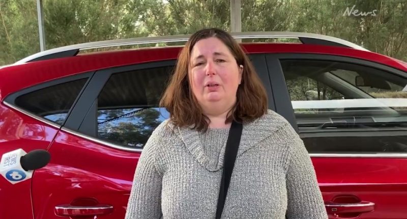 Woman in grey sweater speaks to a news microphone, next to a red car.