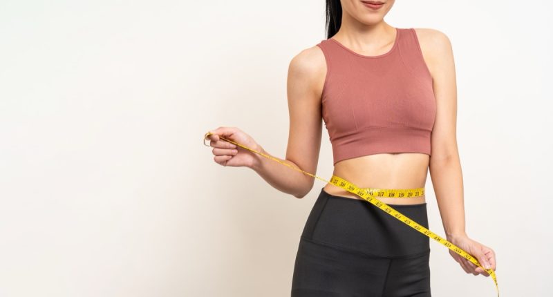 Close up woman measuring her waistline with measure tape. Weight loss diet session. Woman in sportswear slim fit and firm body shape. Care about eating and workout exercise.