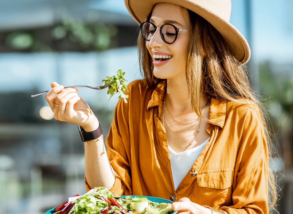 Stylish young woman eating healthy salad on a restaurant terrace, feeling happy on a summer day