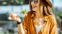 Stylish young woman eating healthy salad on a restaurant terrace, feeling happy on a summer day