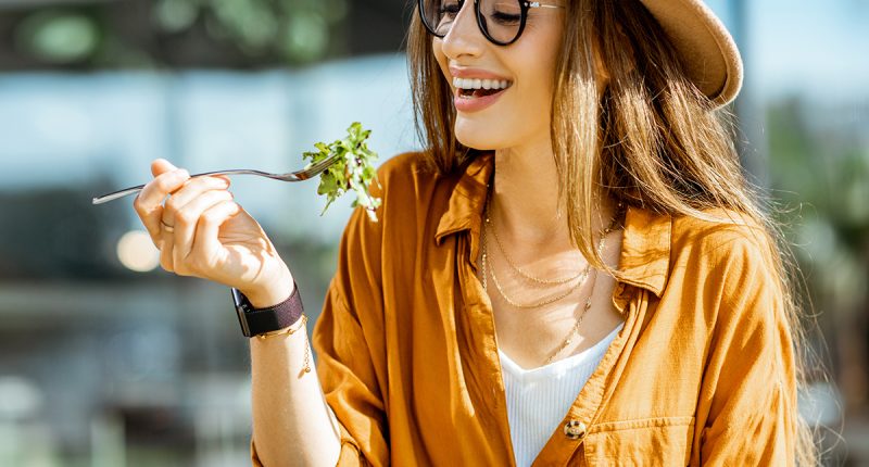 Stylish young woman eating healthy salad on a restaurant terrace, feeling happy on a summer day