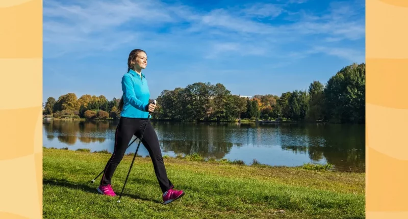 happy young woman doing Nordic walking with walking poles through park by a lake on a sunny day