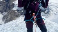 A woman mountain climber poses on a snowy peak, giving a peace sign.