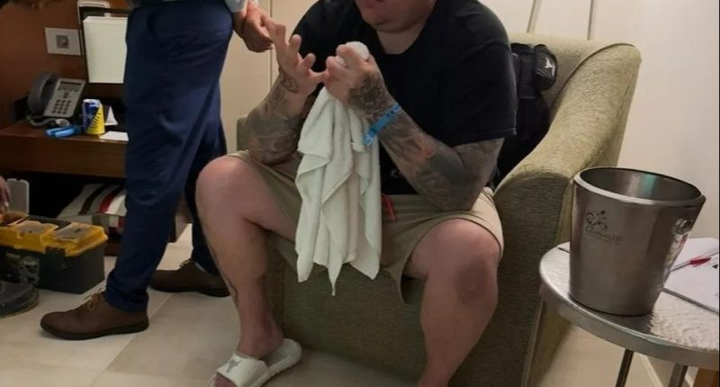 Man seated in a hotel room, holding a towel and looking down at his hands.