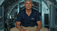 Man in OceanGate shirt sitting in a submersible, holding a controller.
