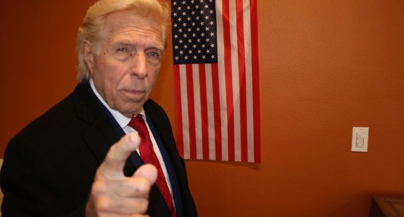 A Donald Trump doppelgänger points at the viewer in front of an American flag.