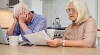 Senior couple reviewing financial documents at their kitchen table, looking distressed.