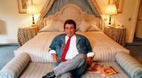 Bob Guccione sitting on a bed in a denim jacket and red tie, holding magazines.