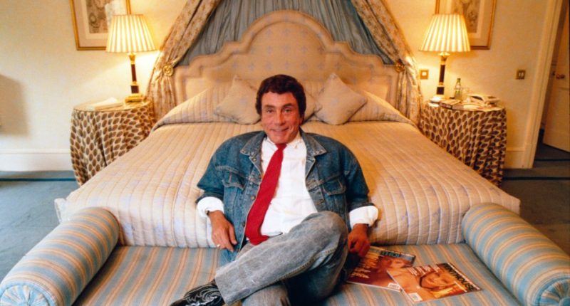 Bob Guccione sitting on a bed in a denim jacket and red tie, holding magazines.