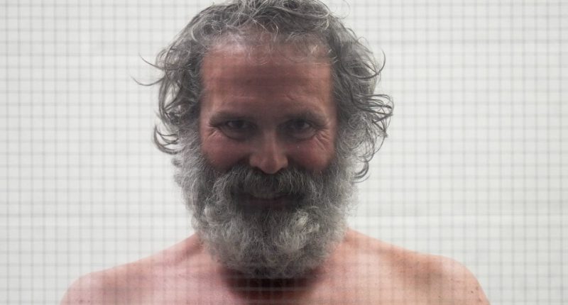 Shirtless man with long gray hair and beard behind glass.