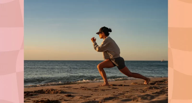 fit woman in biker shorts, a sweatshirt, and a baseball cap doing walking lunges on the beach at sunset