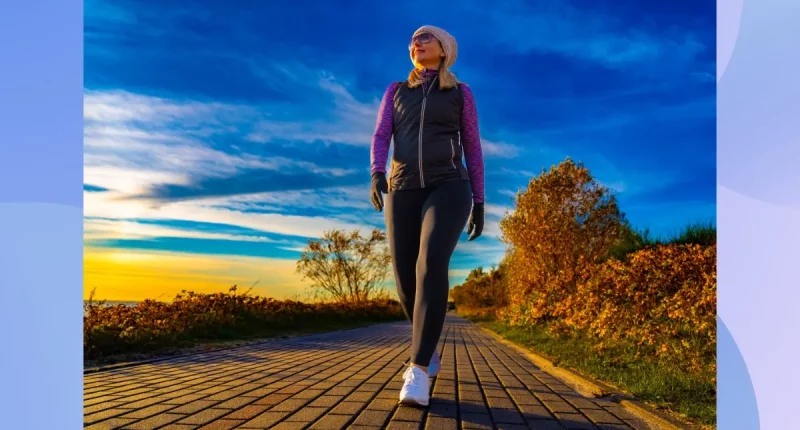 fit, middle-aged woman bundled up and walking on path by the water on fall afternoon at sunset