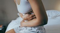 Menstruation, sick and stomach ache with black woman in bedroom for indigestion, cramps and illness. Frustrated, gas and stress with girl on bed for constipation, bloating and intestine problems