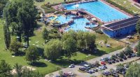 Aerial view of an outdoor pool complex with a water slide, surrounded by a grassy area with people sunbathing and a parking lot.