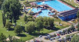 Aerial view of an outdoor pool complex with a water slide, surrounded by a grassy area with people sunbathing and a parking lot.