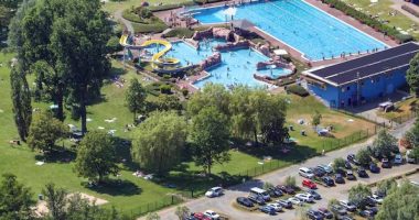 Aerial view of an outdoor pool complex with a water slide, surrounded by a grassy area with people sunbathing and a parking lot.