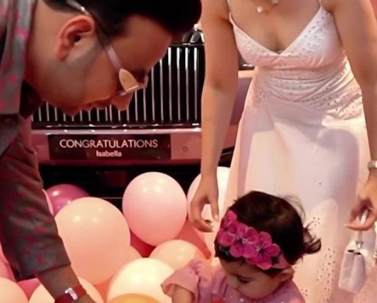 A toddler girl receives a car key from her parents in front of a pink Rolls Royce.