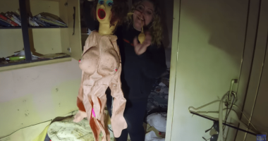 Woman holding up a sex doll in a dilapidated room.