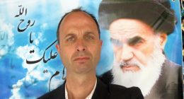 A man in a suit posing with a large photo of Ayatollah Khomeini in the background.