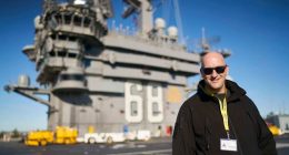 Man in sunglasses standing on the deck of an aircraft carrier.