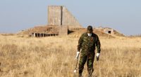 A man in camouflage clothing measures radiation levels near the remains of a structure.