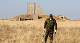 A man in camouflage clothing measures radiation levels near the remains of a structure.